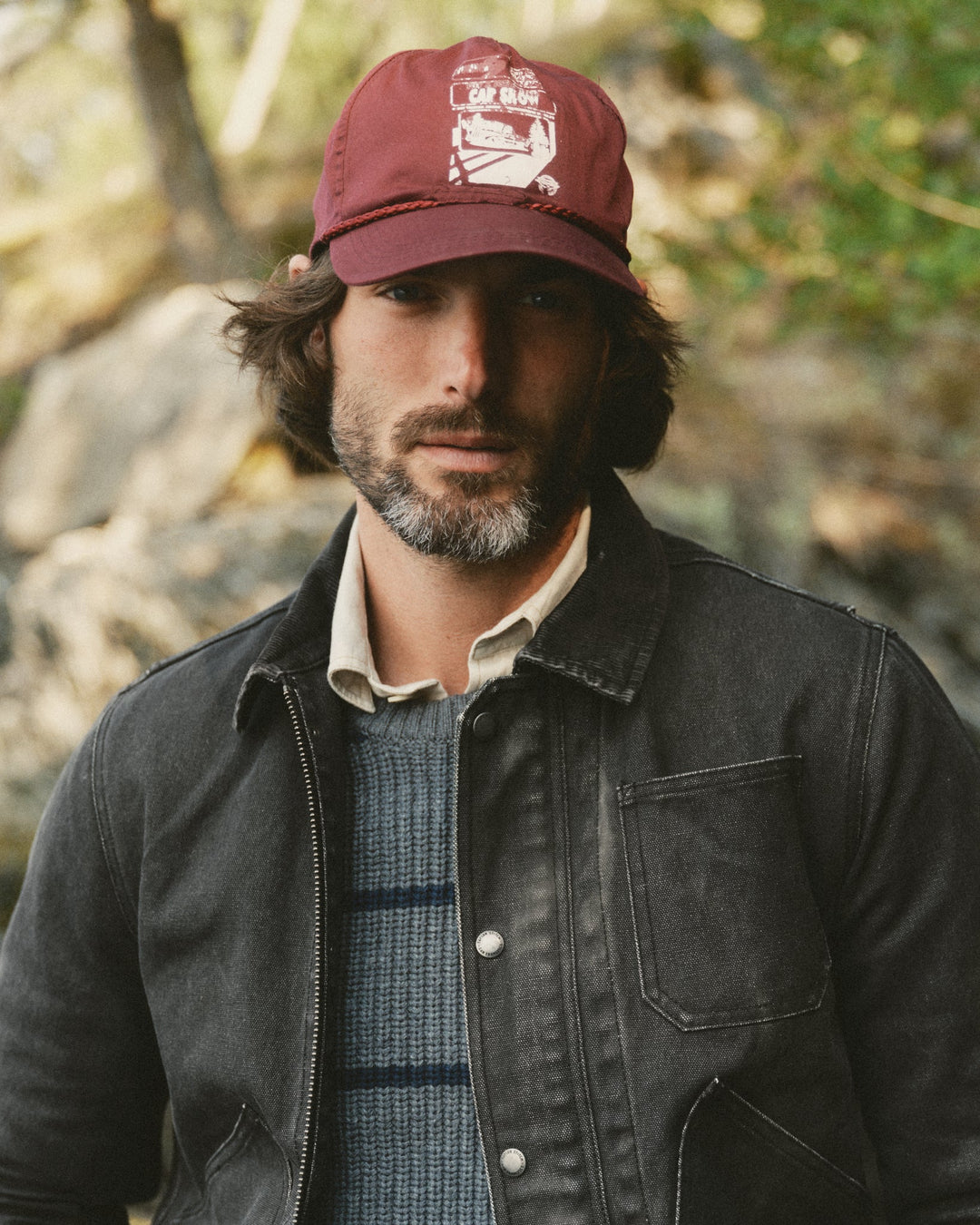 A man wearing a dark charcoal gray canvas utility jacket over a blue-gray knit sweater, a cream collared shirt, and a maroon baseball cap with a white graphic, set against a blurred outdoor background of trees and rocks.