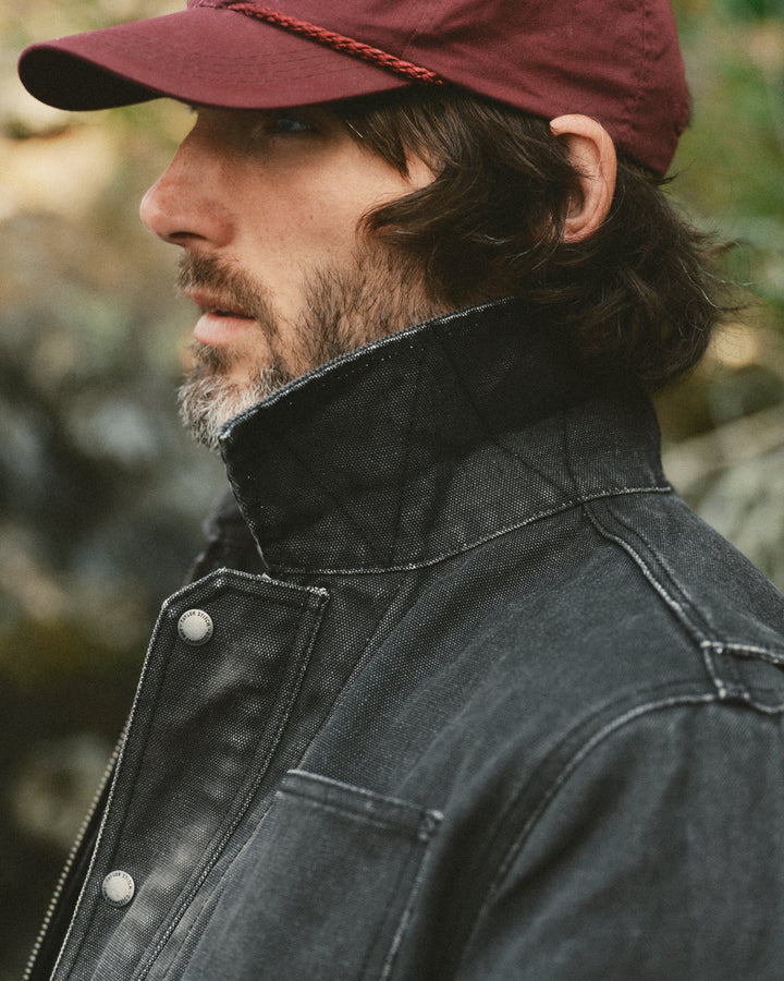 A close-up side profile of a man wearing a dark charcoal gray high-collar denim jacket and a maroon baseball cap with a braided cord detail on the visor, set against a blurred outdoor background.