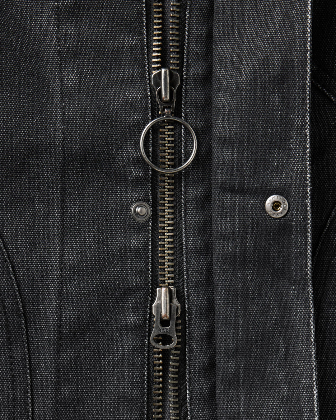 Close-up of a black textured canvas jacket featuring a central silver metal zipper with a circular ring pull, a silver rivet, and a matching zipper pull at the bottom.