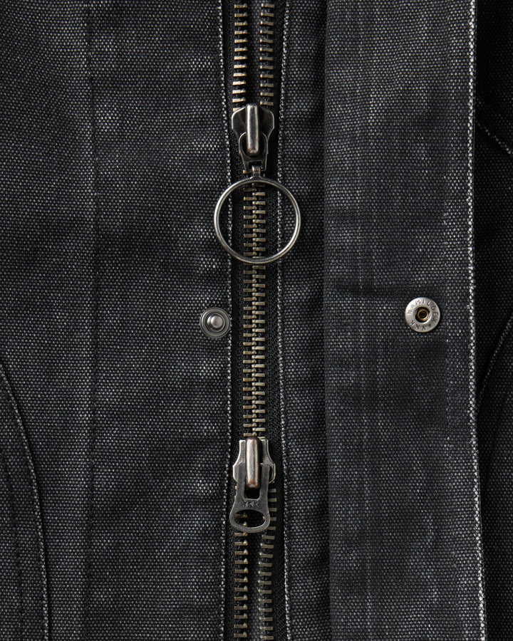 Close-up of a black textured canvas jacket featuring a central silver metal zipper with a circular ring pull, a silver rivet, and a matching zipper pull at the bottom.