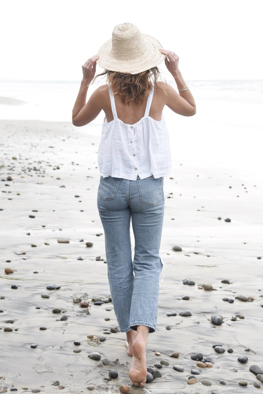 Woman walking on a pebbly beach wearing a white sleeveless top and blue jeans.