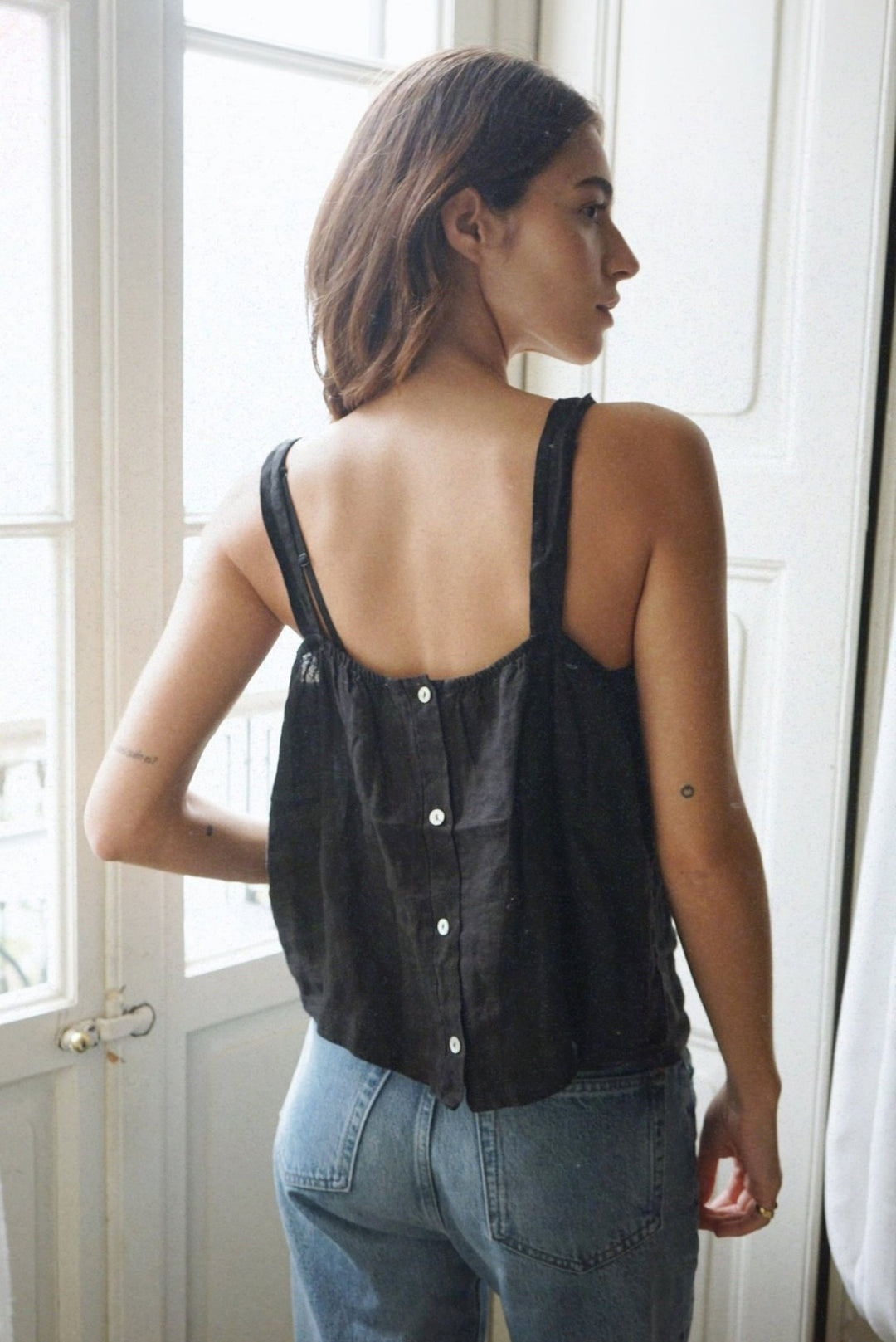 Woman wearing a black sleeveless top with buttons and blue jeans, standing in a room with white walls and a window.