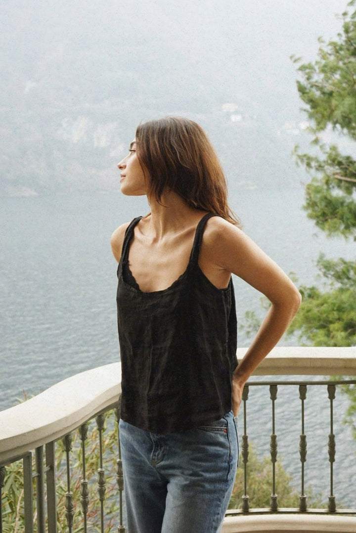 Woman wearing a black tank top and blue jeans standing on a balcony with a scenic view.