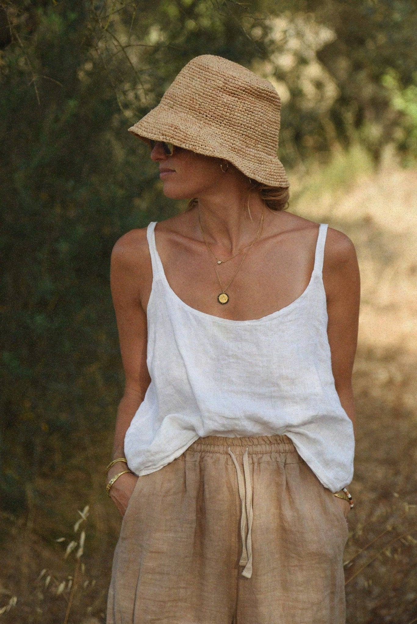 Woman wearing a white tank top and beige pants with a straw hat in a natural setting.