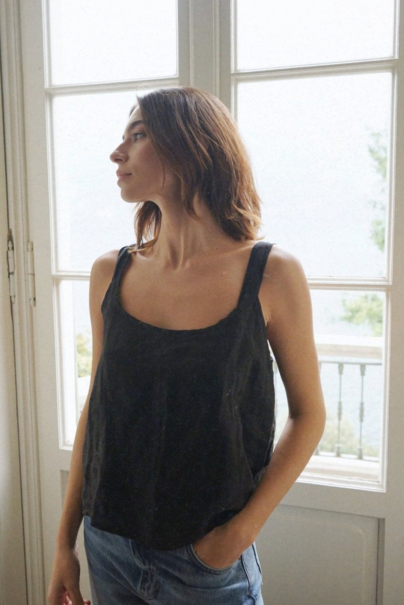 Woman wearing a black tank top and blue jeans standing in a room with large windows.