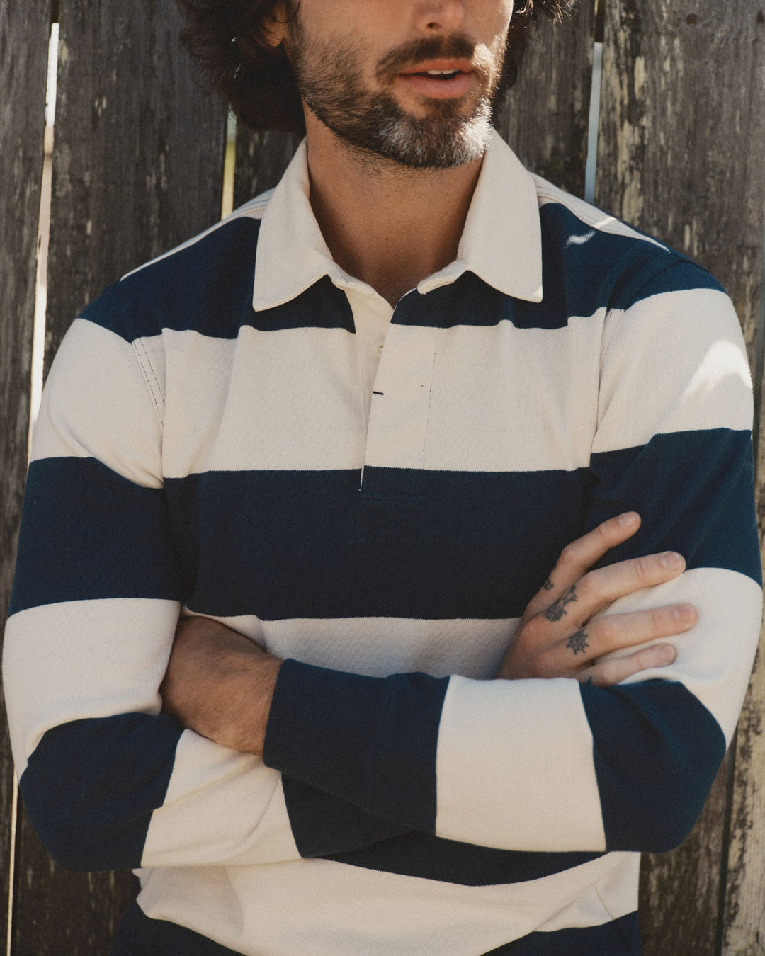A man wearing a long-sleeve rugby-style shirt with navy blue and off-white horizontal stripes, standing with arms crossed against a weathered wooden fence.