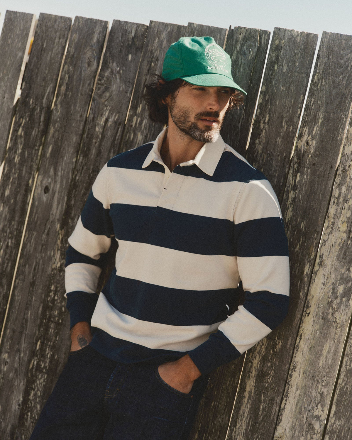 A man wearing a green baseball cap and a navy and cream horizontally striped long-sleeve rugby shirt leans against a weathered wooden fence.