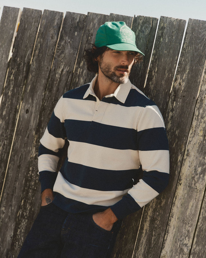A man wearing a green baseball cap and a navy and cream horizontally striped long-sleeve rugby shirt leans against a weathered wooden fence.