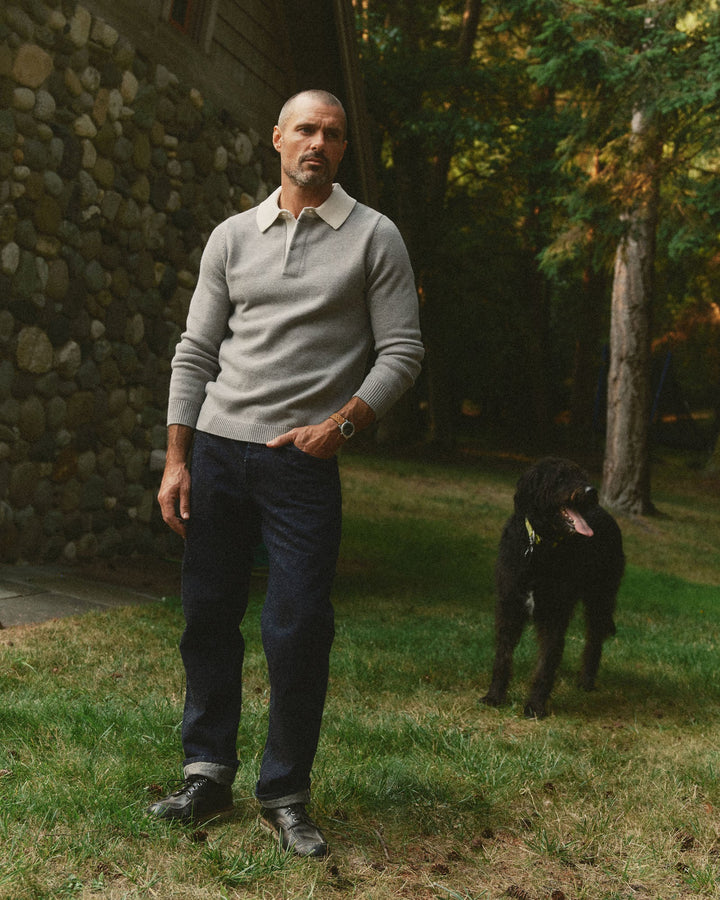 A man wearing a light gray long-sleeve rugby sweater, dark blue jeans, and black shoes stands on a grassy lawn next to a stone wall, with a black dog in the background among trees.
