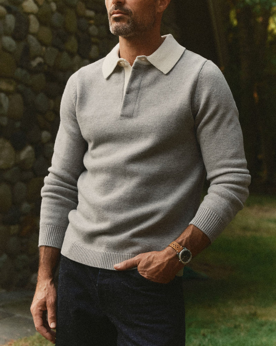 A man wearing a light gray knit rugby sweater with a contrasting cream collar, paired with dark jeans and a wooden watch, standing outdoors against a stone wall.