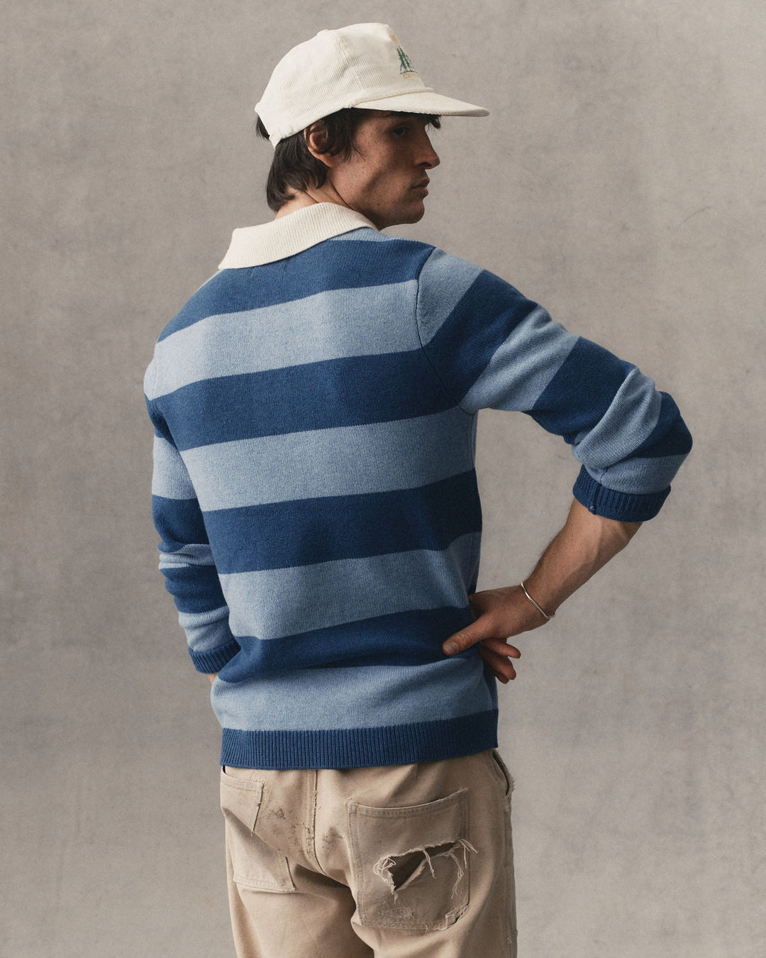 A man wearing a blue and light blue horizontally striped knit sweater, a cream baseball cap, and beige pants, photographed against a textured gray wall.