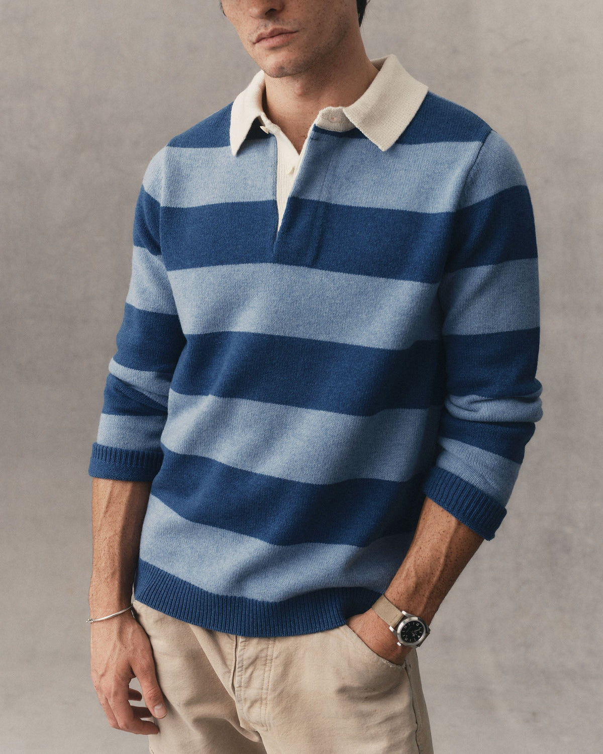 A man wearing a blue and light blue horizontally striped knit rugby sweater with a cream collar, paired with beige trousers, against a textured gray background.
