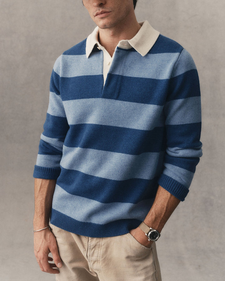 A man wearing a blue and light blue horizontally striped knit rugby sweater with a cream collar, paired with beige trousers, against a textured gray background.