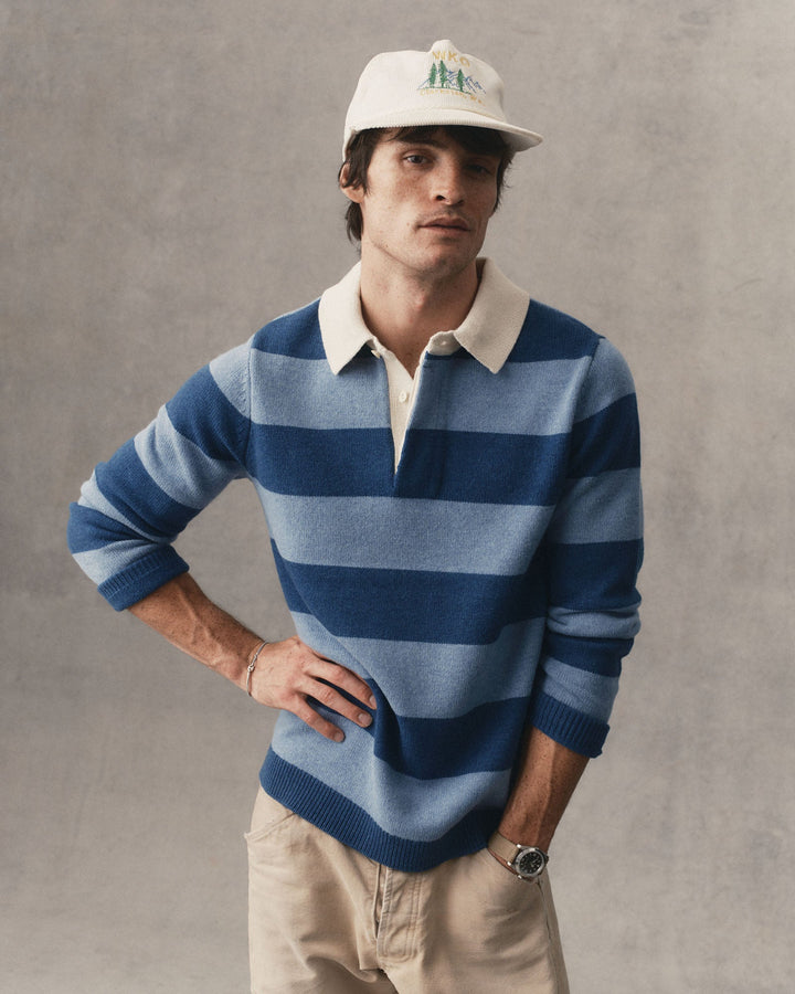 A man wearing a blue and light blue horizontally striped knit rugby sweater with a cream collar, paired with beige pants and a cream baseball cap, standing against a textured gray wall.