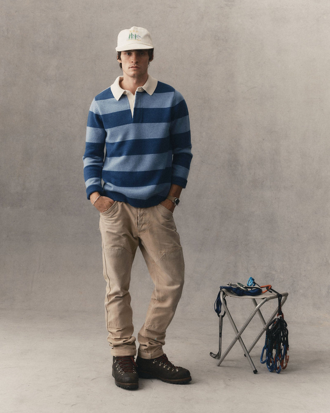 A man stands against a textured gray studio wall wearing a blue and navy striped long-sleeve rugby sweater, beige trousers, and dark brown hiking boots. He is also wearing a white cap.
