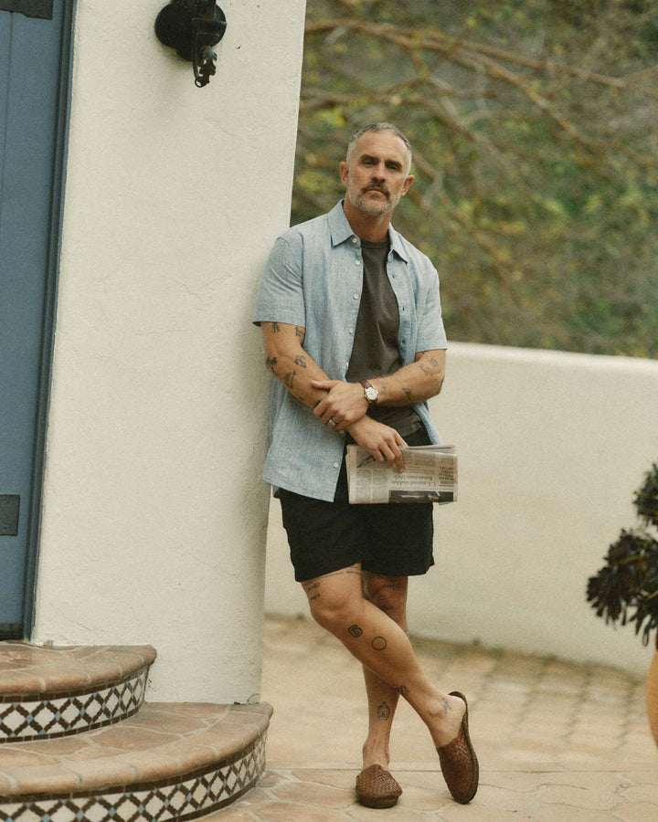 Man standing outdoors holding a newspaper, wearing The Short Sleeve California in Sky Hempand black shorts.