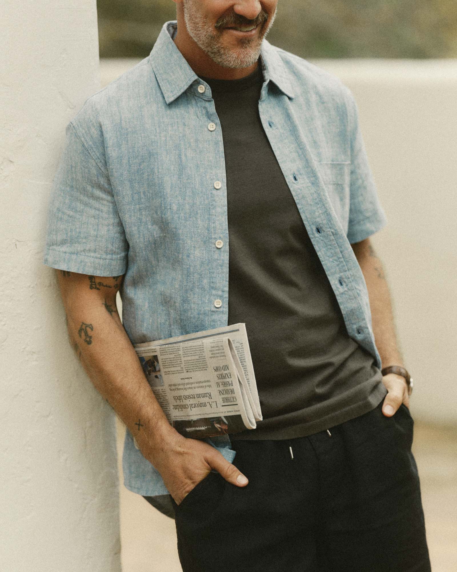 Man wearing The Short Sleeve California in Sky Hempover a black t-shirt, holding a newspaper, against a neutral background.