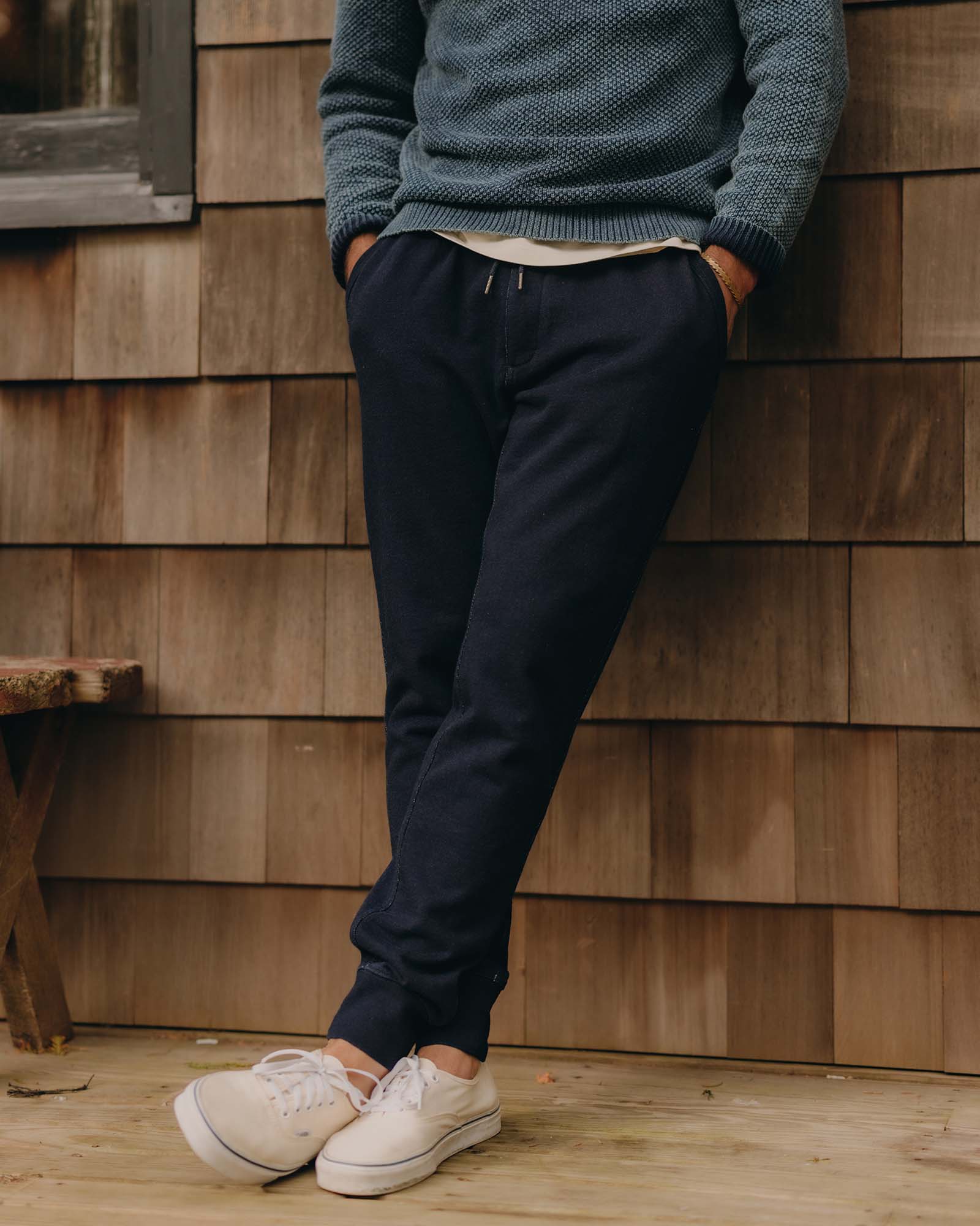 Person wearing a blue sweater and Taylor Stitch's Sunset Pant in Rinsed Indigo Terry leaning against a wooden wall.