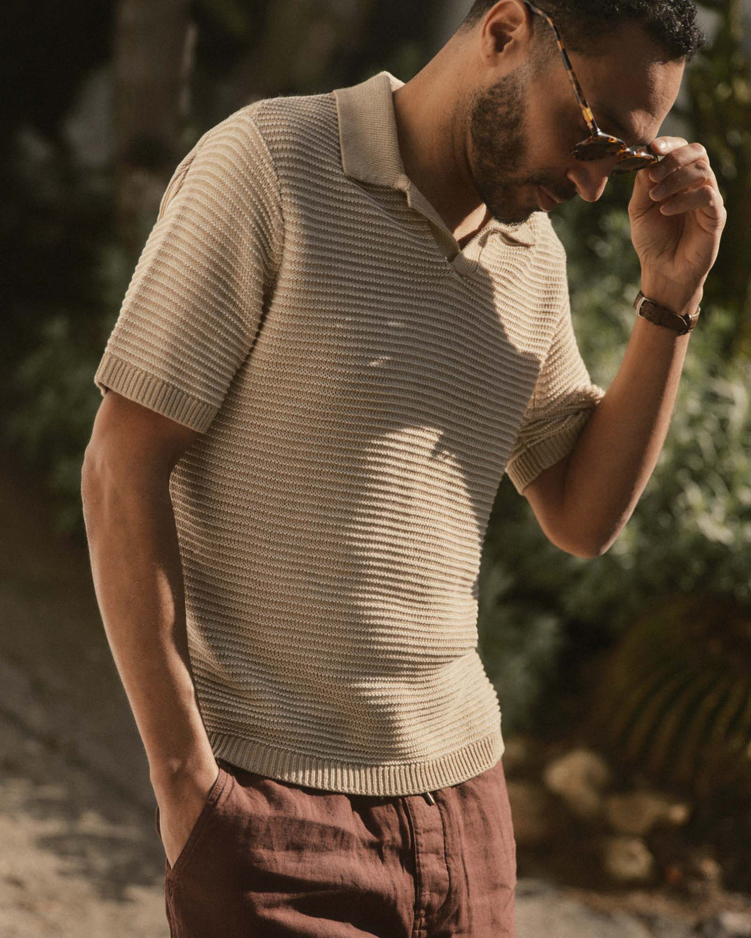 Man wearing The Valencia Sweater Polo
in Sand Stripe and sunglasses, standing outdoors with greenery in the background.