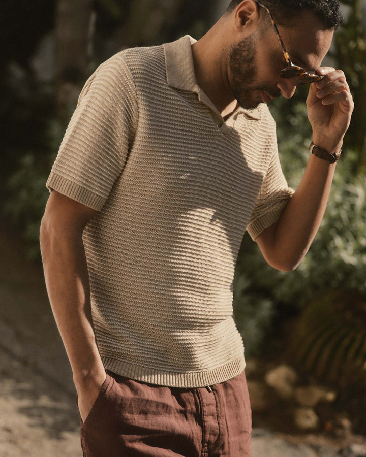 Man wearing The Valencia Sweater Polo
in Sand Stripe and sunglasses, standing outdoors with greenery in the background.