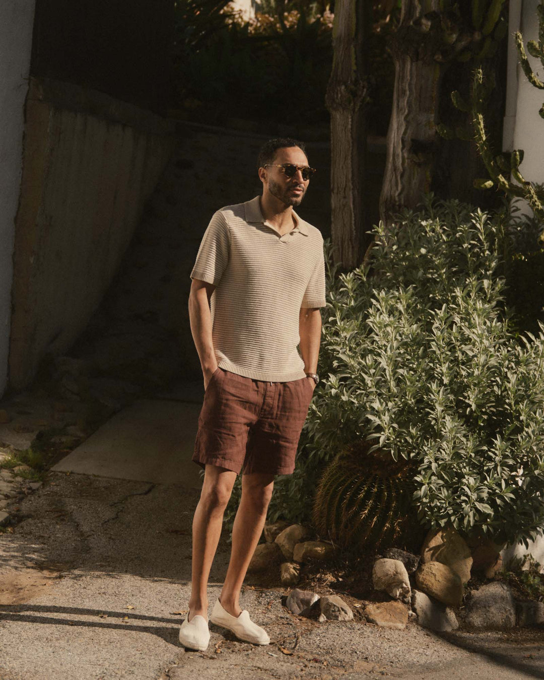 Man in The Valencia Sweater Polo
in Sand Stripe and brown shorts standing outdoors near a building and plants
