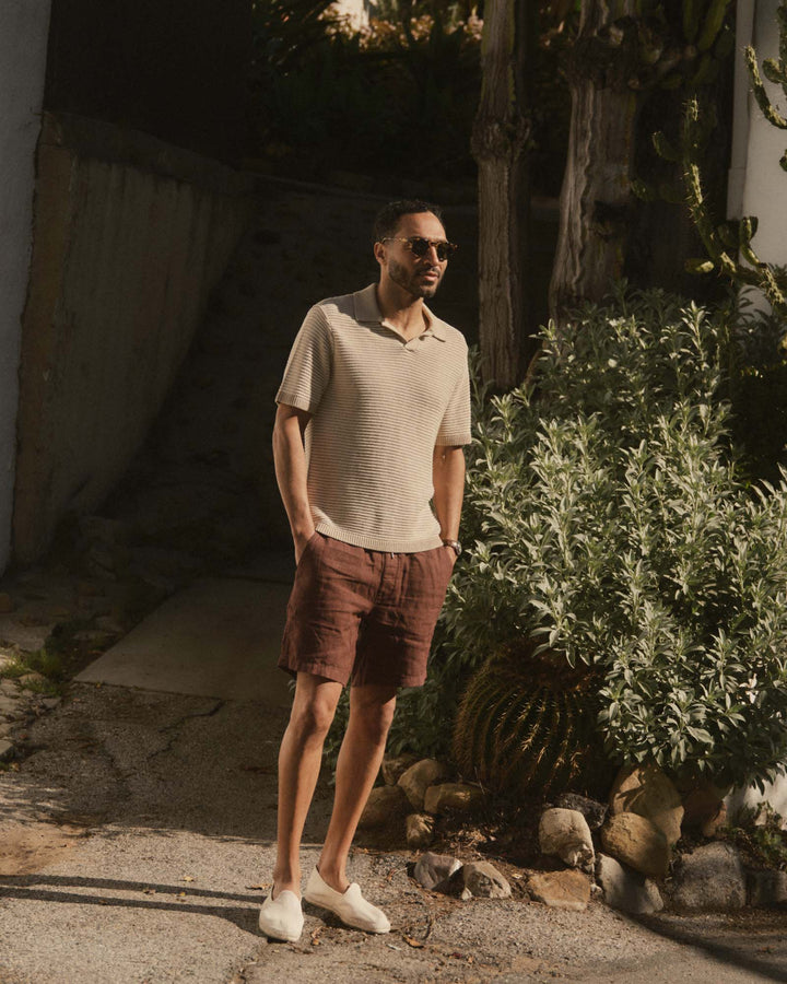 Man in The Valencia Sweater Polo
in Sand Stripe and brown shorts standing outdoors near a building and plants