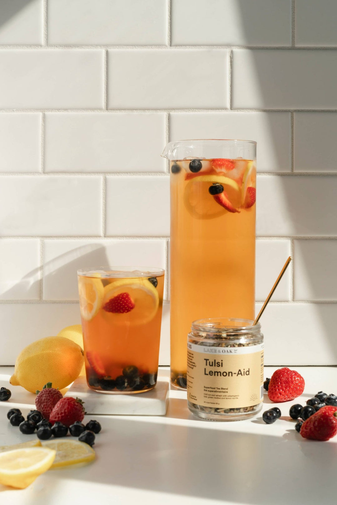 Two glasses of iced tea with fruit and a jar of Tulsi Lemon Aid on a white tiled wall background.