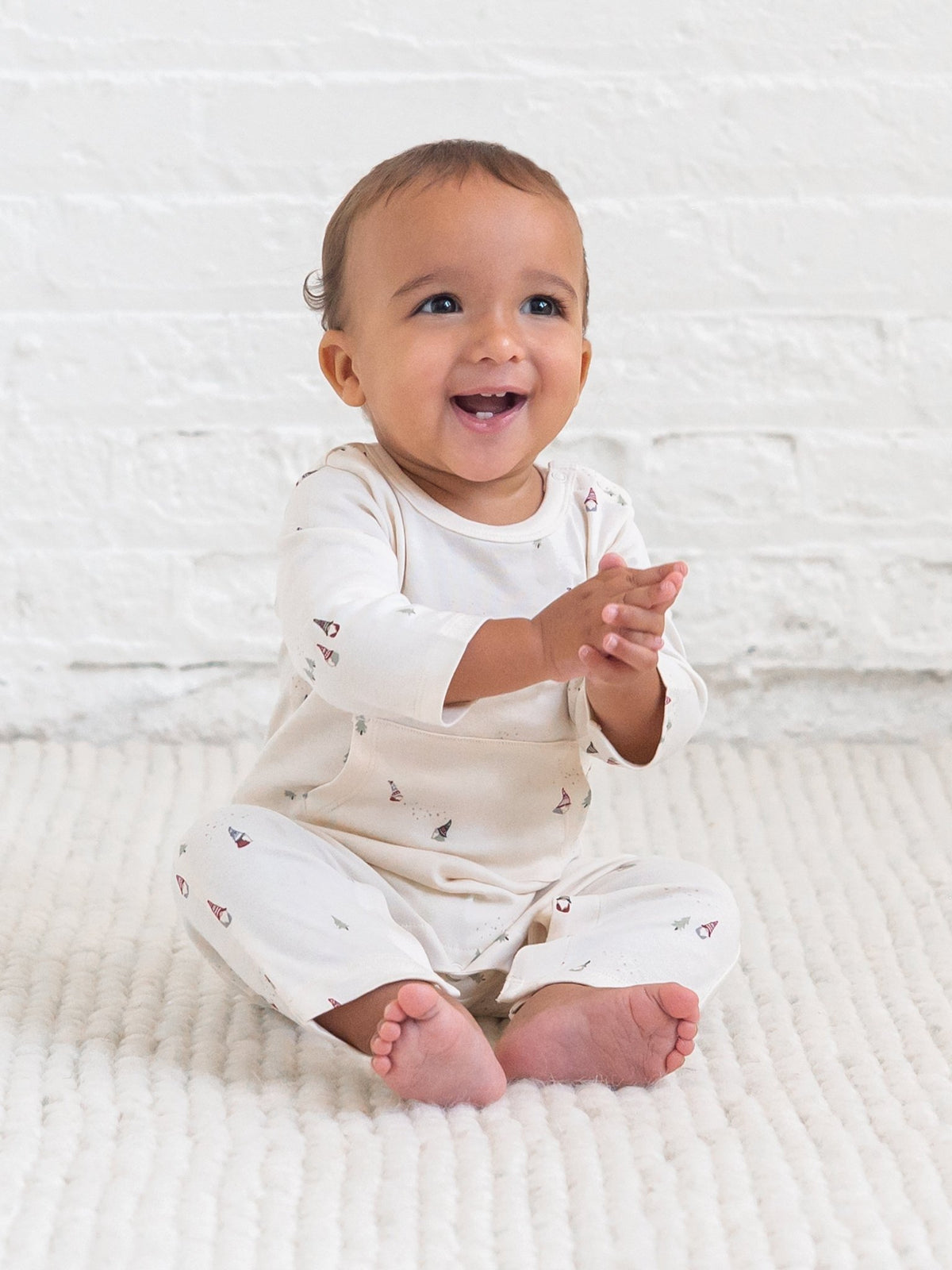 A smiling baby sitting on a textured cream blanket in front of a white brick wall, wearing a Colored Organics' gnome print Una Pocket Romper.