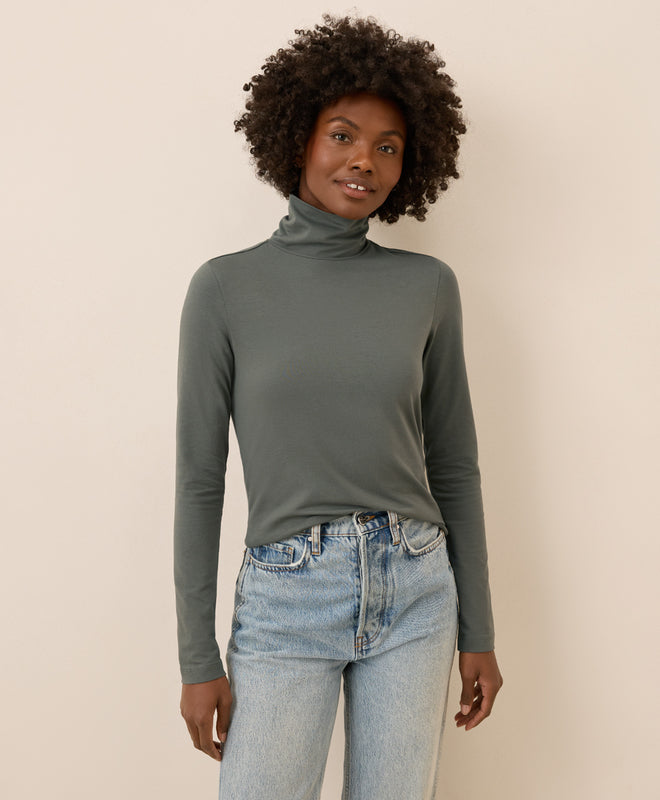 Woman wearing a castor green turtleneck and light blue jeans against a beige background