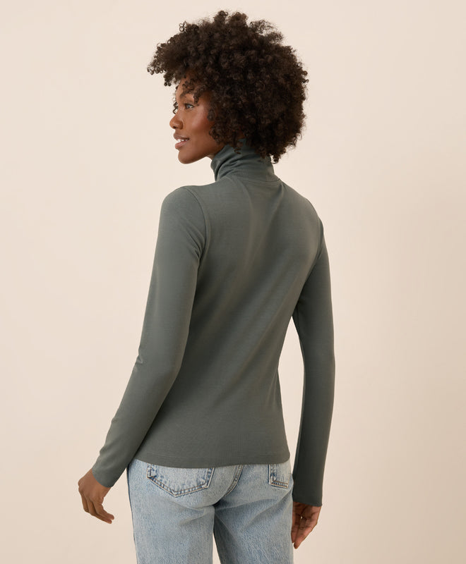Woman wearing a castor green turtleneck sweater and light blue jeans against a beige background