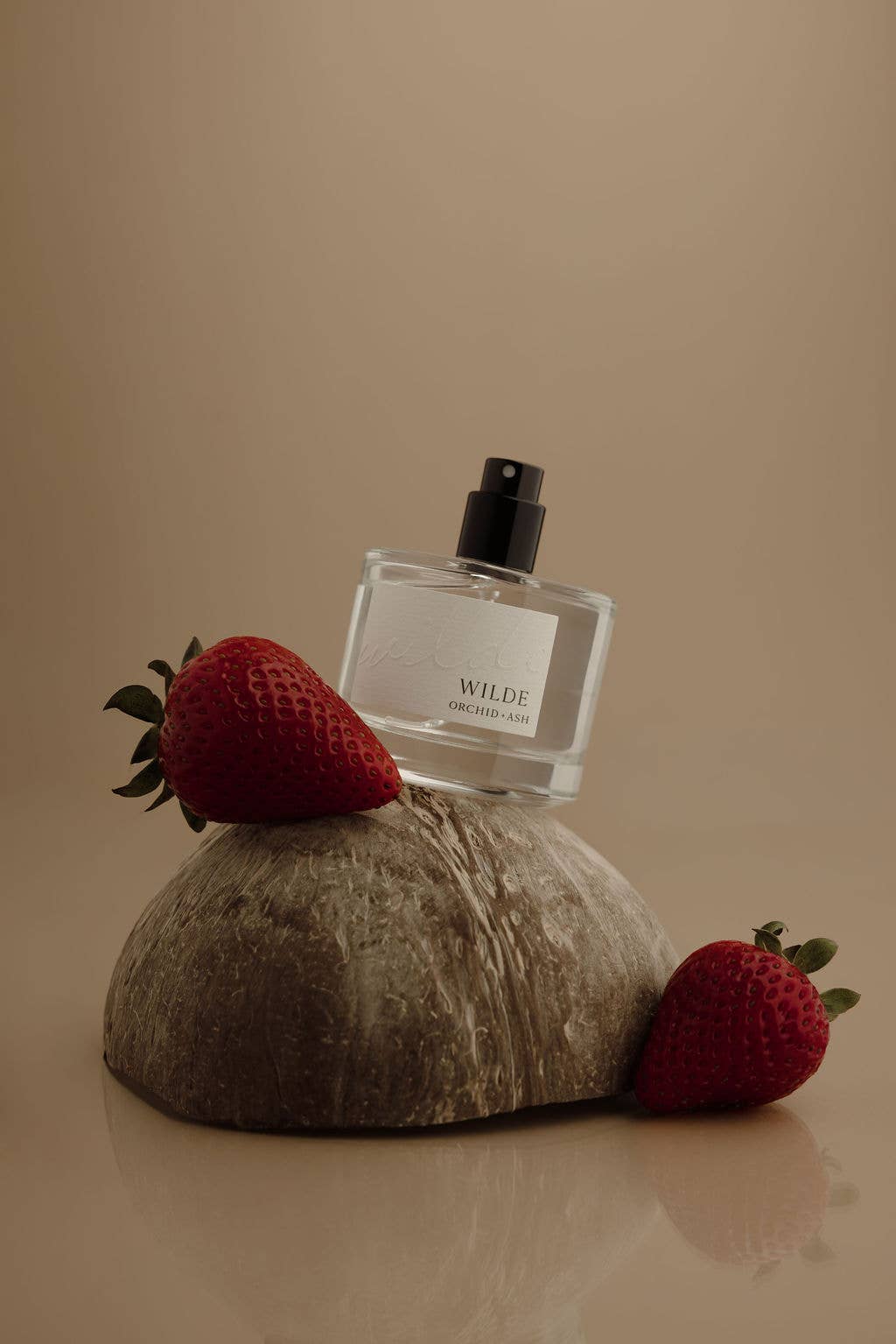 A clear rectangular perfume bottle with a black spray top and a white label reading WILDE, styled on a textured stone slab with two red strawberries on a warm beige background.