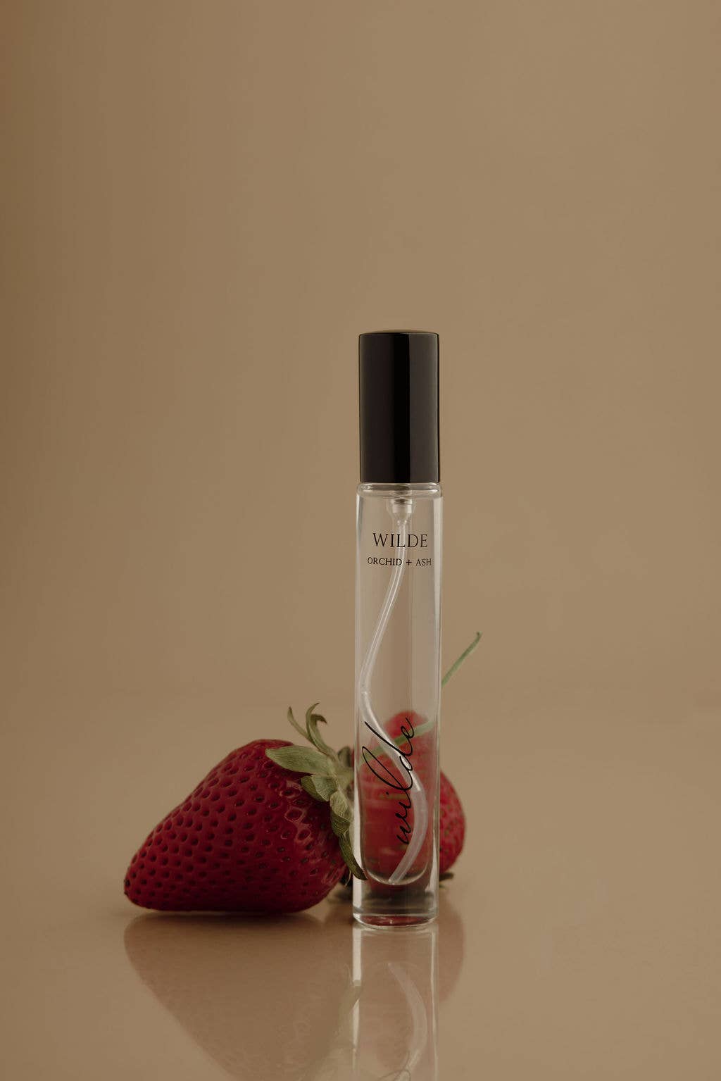 WILDE Perfume Travel Spray with a black cap stands on a beige surface against a warm beige background, styled with two red strawberries in front. The bottle label reads WILDE Orchid + Ash.