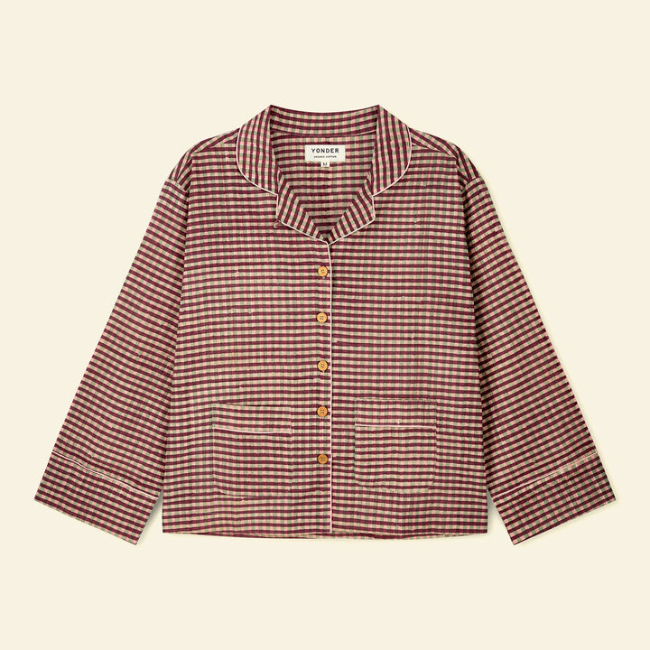 A long-sleeve wood button-front pajama top with a small-scale checkered pattern in burgundy and beige, featuring a collar and front pockets, laid flat on a light cream background.