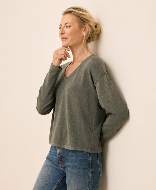 Woman wearing a green sweater and blue jeans against a beige wall