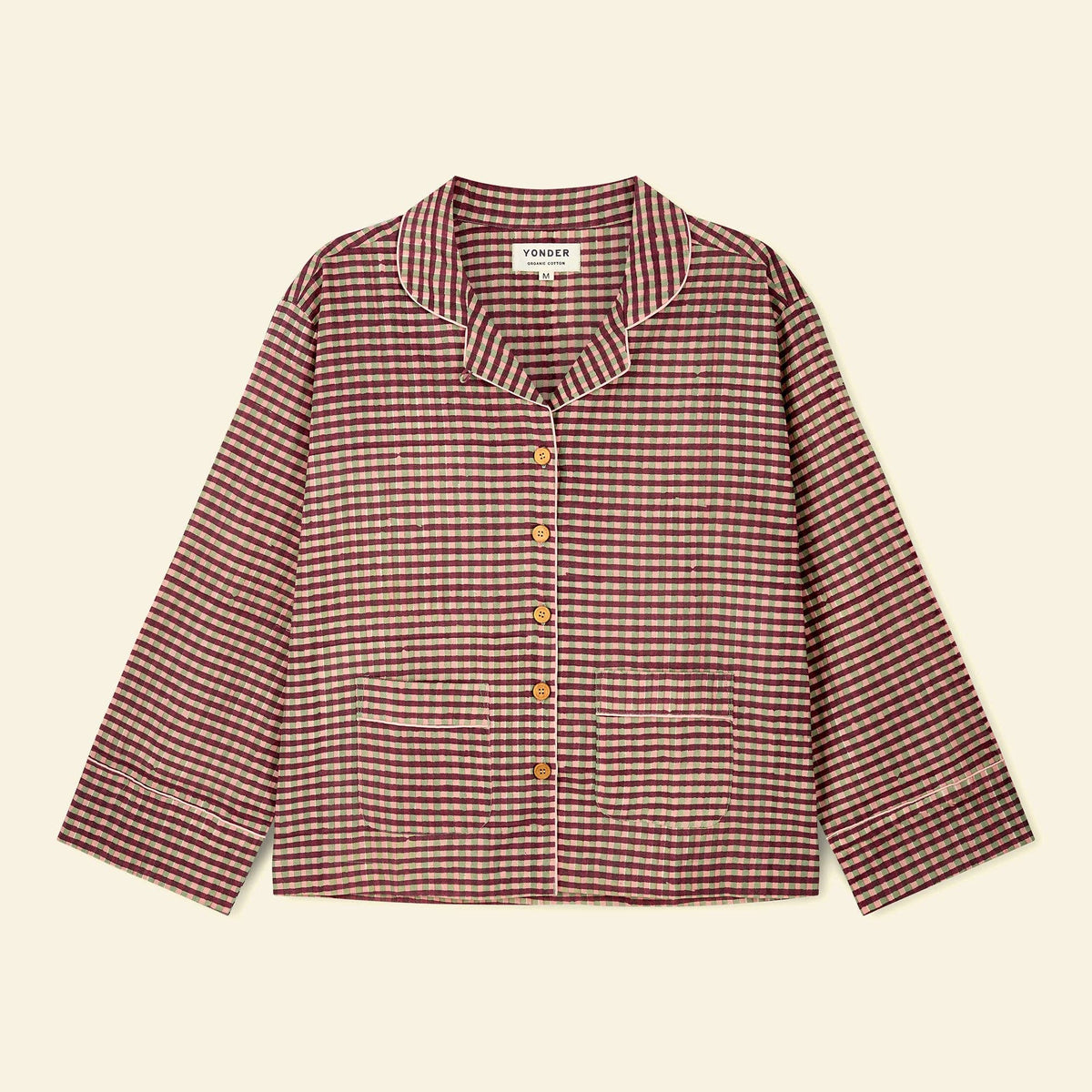 A long-sleeve wood button-front pajama top with a small-scale checkered pattern in burgundy and beige, featuring a collar and front pockets, laid flat on a light cream background.